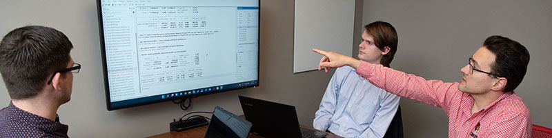 Three students sit at a table looking at a TV screen of information.