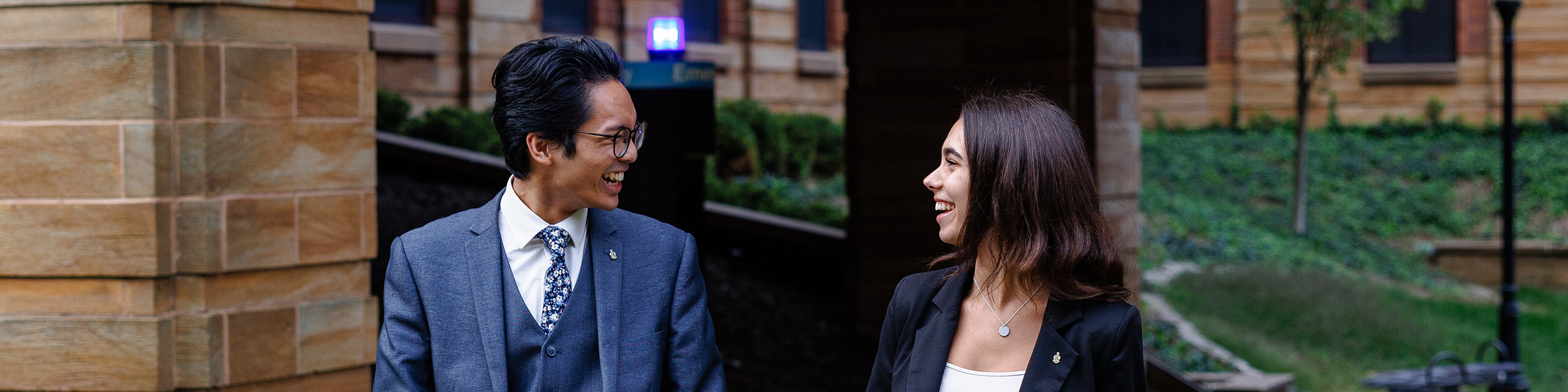 A man and woman smiling at each other in front of a brick building and greenery.