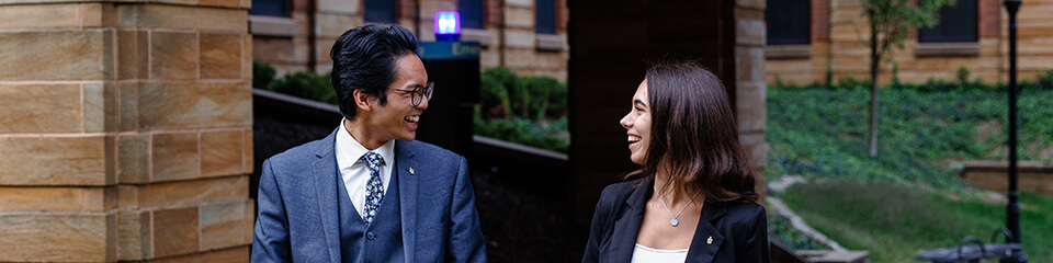 A man and woman smiling at each other in front of a brick building and greenery.